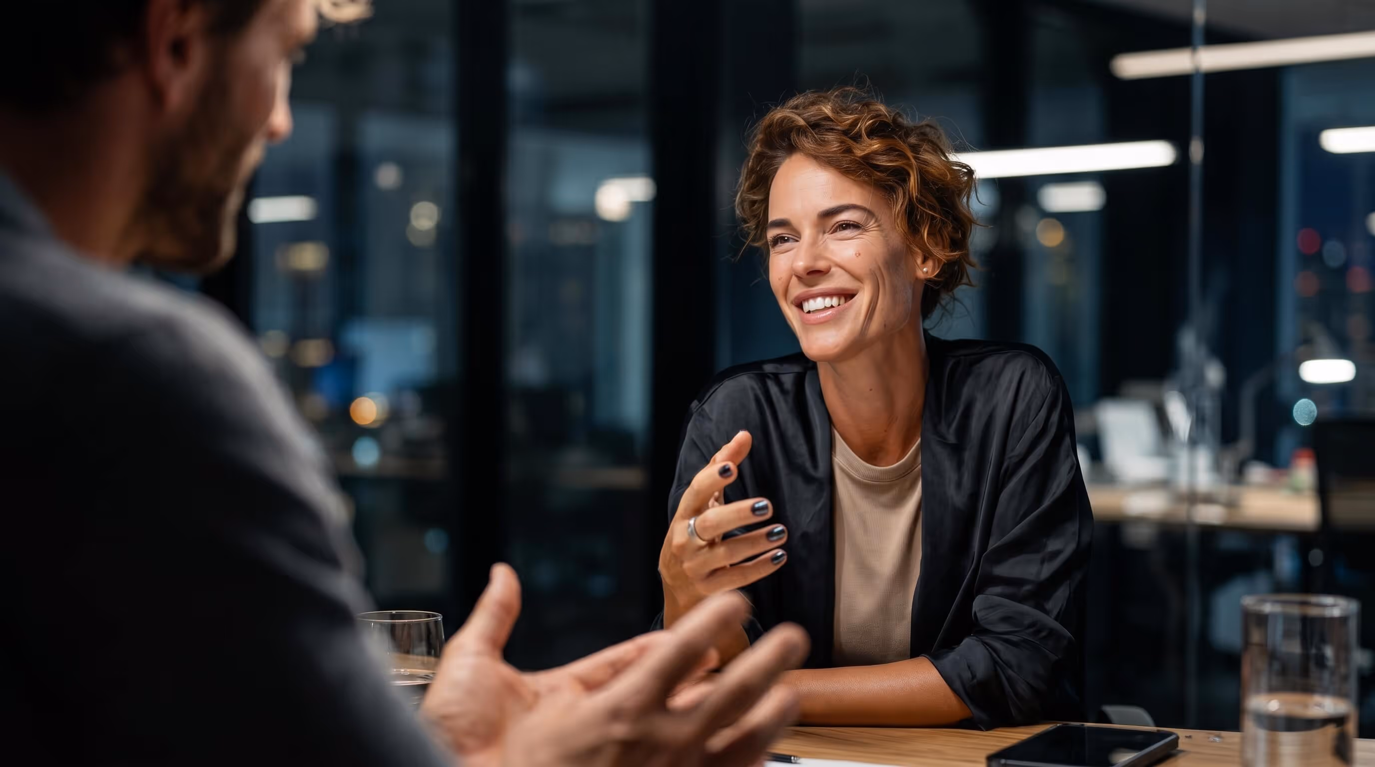 Smiling woman with curly hair engaged in a conversation with a man in a modern office setting.